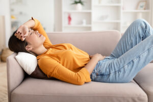 Young asian woman lying on couch with one hand on forehead and one hand on low abdomen, as if from menstrual cramps
