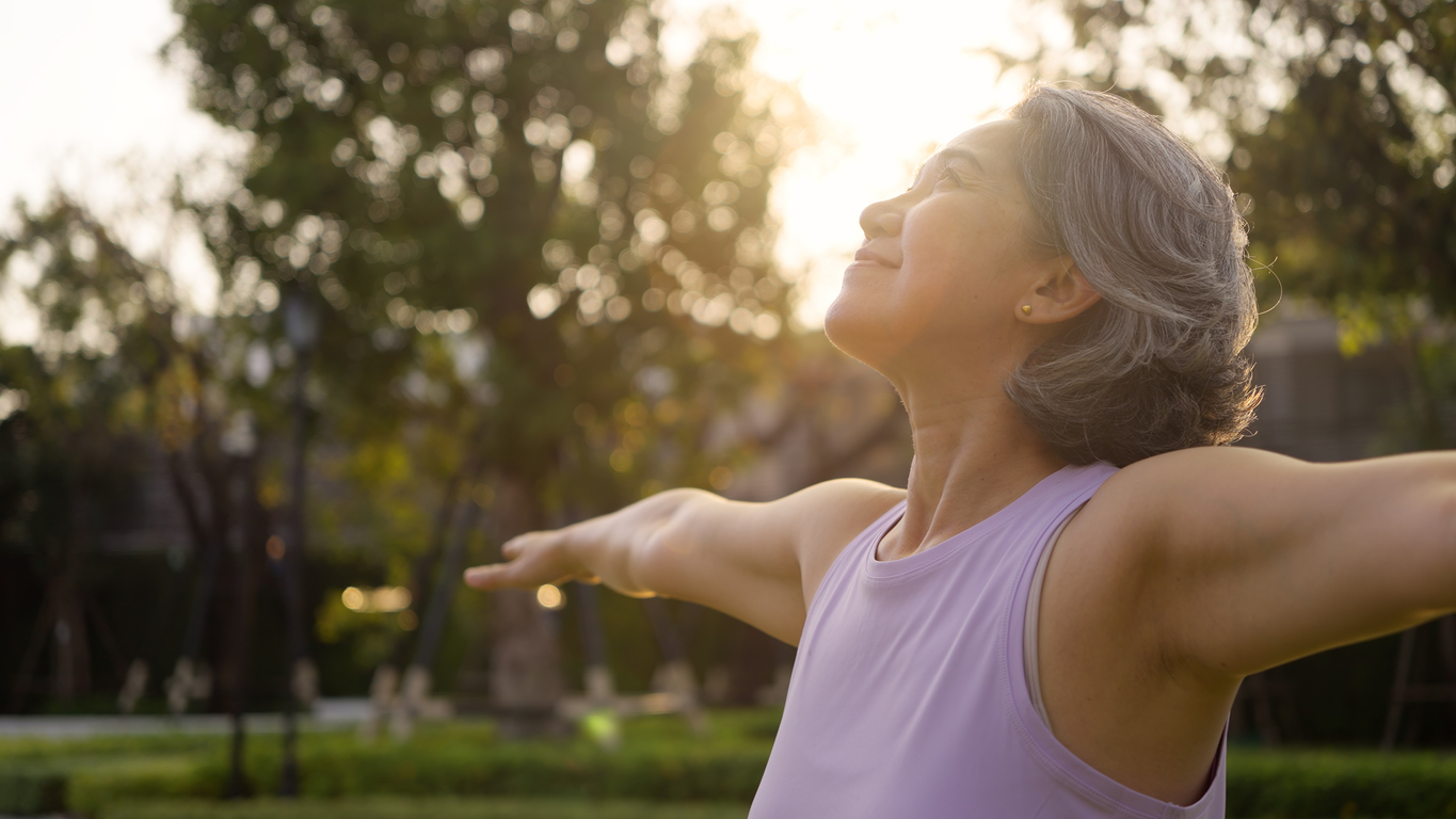 Mature asian woman with arms outstretched in a park, in golden sunlight-healthy aging concept