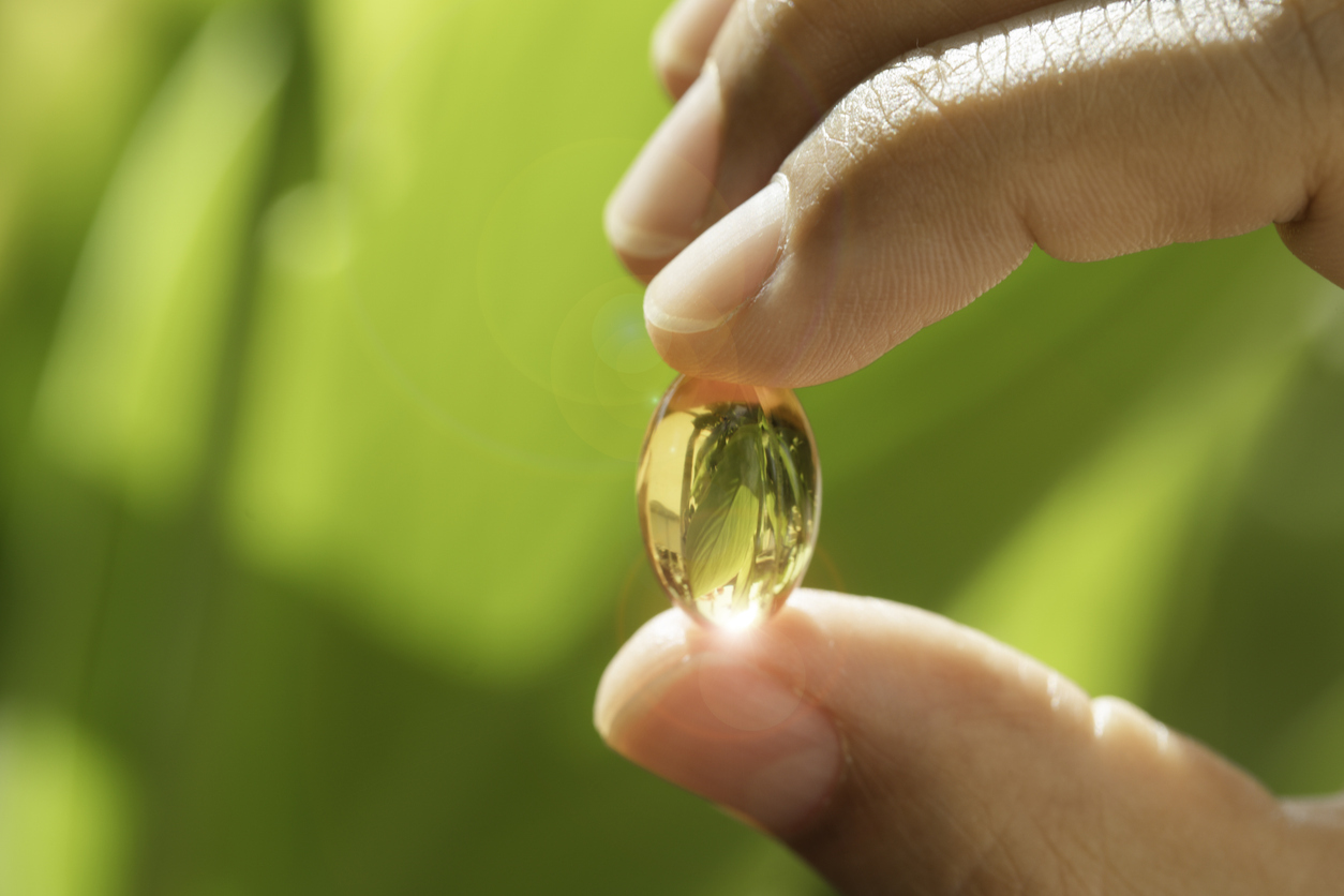 Close up of hand showing small gel capsule, which could be vitamin E, green, plant background